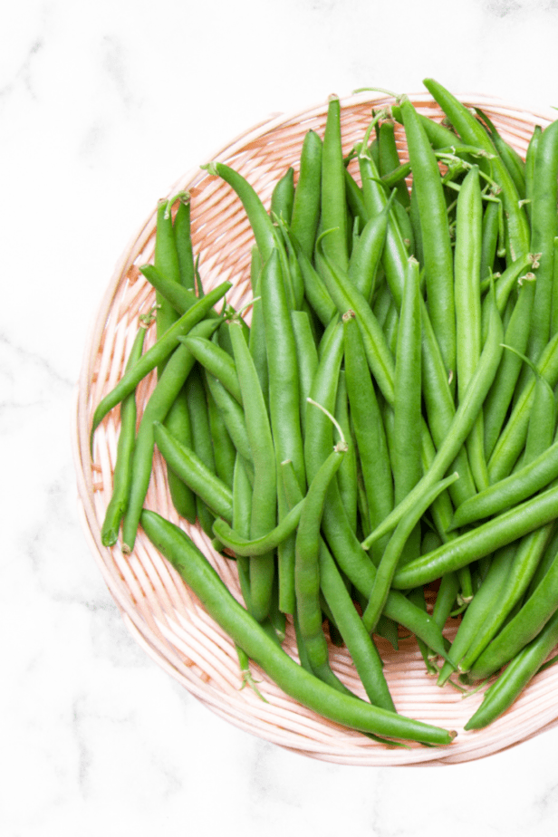 a bunch of green beans in a wood woven basket on a white marble counter